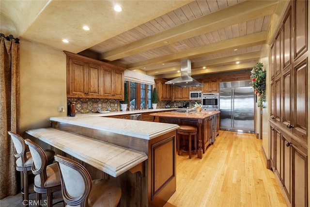 kitchen featuring backsplash, kitchen peninsula, built in appliances, a kitchen island with sink, and island range hood