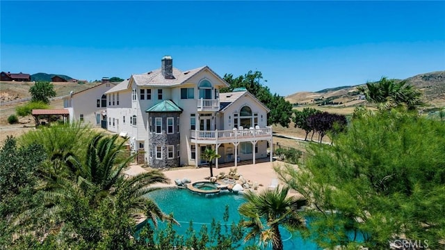 rear view of house featuring a patio, a chimney, a pool with connected hot tub, a mountain view, and a balcony