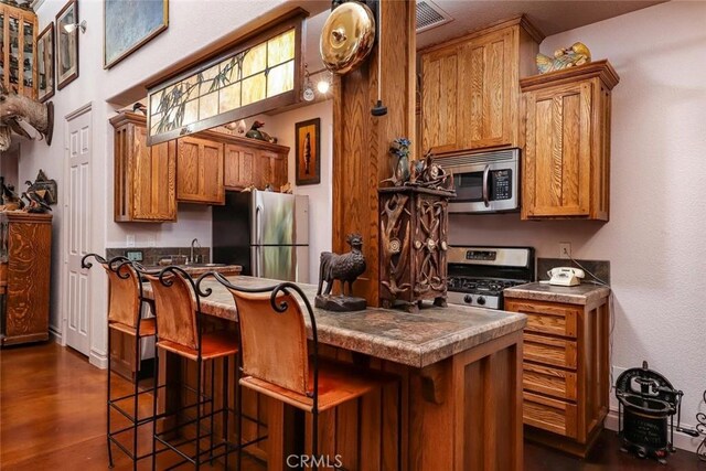 kitchen with a sink, appliances with stainless steel finishes, brown cabinets, dark wood-style floors, and a kitchen bar