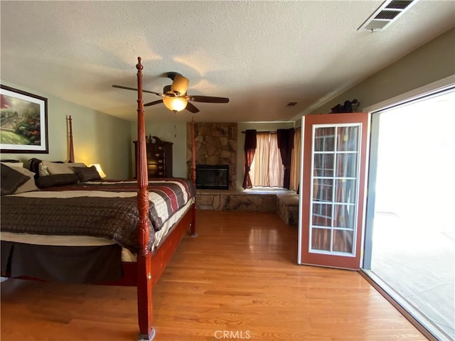 bedroom with visible vents, light wood-style flooring, ceiling fan, a stone fireplace, and a textured ceiling