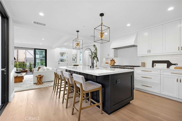 kitchen featuring pendant lighting, white cabinets, premium range hood, light wood-type flooring, and a center island with sink