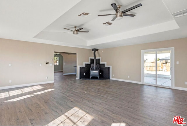 unfurnished living room with ceiling fan, a wood stove, a tray ceiling, and dark hardwood / wood-style floors