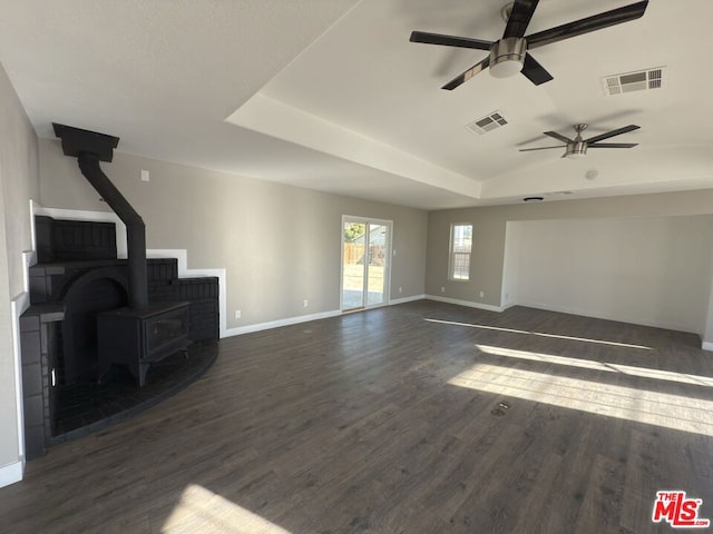 unfurnished living room featuring a raised ceiling, ceiling fan, dark wood-type flooring, and a wood stove