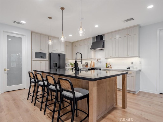 kitchen featuring black fridge with ice dispenser, sink, a center island with sink, light wood-type flooring, and wall chimney range hood