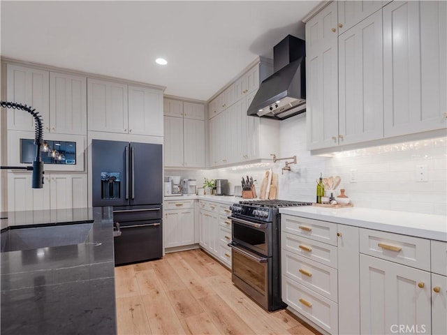 kitchen featuring high end black refrigerator, tasteful backsplash, range with two ovens, wall chimney exhaust hood, and light hardwood / wood-style flooring