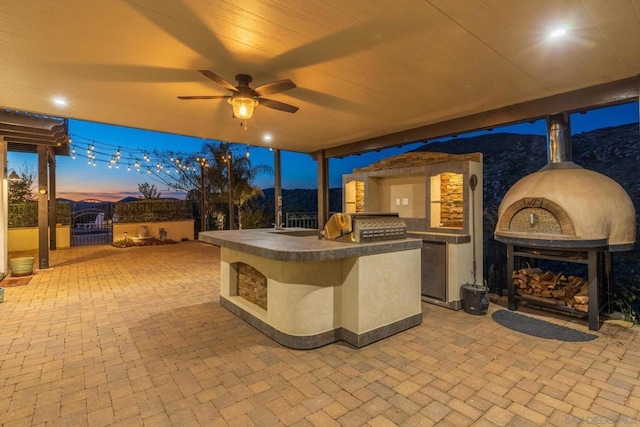 patio terrace at dusk featuring exterior kitchen, an outdoor fireplace, and ceiling fan