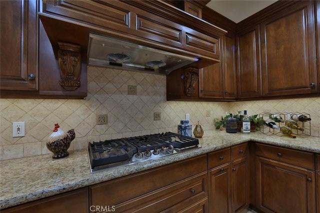 kitchen with ventilation hood, decorative backsplash, stainless steel gas cooktop, dark brown cabinets, and light stone counters