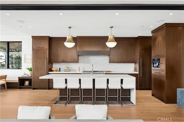 kitchen with decorative light fixtures, tasteful backsplash, light hardwood / wood-style floors, custom exhaust hood, and a center island with sink