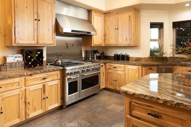 kitchen with range with two ovens, stone finish flooring, light brown cabinetry, dark stone counters, and wall chimney exhaust hood