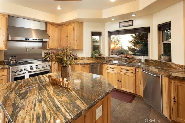 kitchen with recessed lighting, stainless steel appliances, a sink, wall chimney range hood, and dark stone countertops