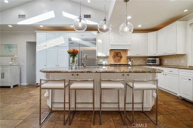 kitchen featuring stone countertops, stone tile floors, stainless steel built in refrigerator, a kitchen bar, and an island with sink