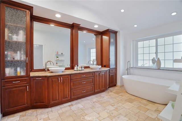 full bathroom with stone tile floors, double vanity, recessed lighting, a freestanding bath, and a sink