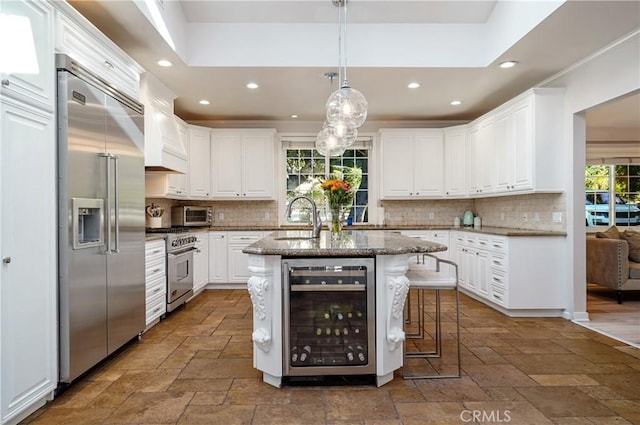 kitchen with premium appliances, stone tile floors, a kitchen island with sink, white cabinetry, and beverage cooler