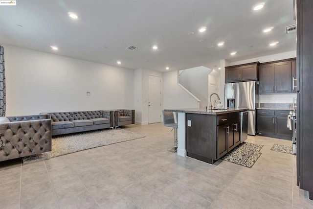 kitchen featuring stainless steel fridge with ice dispenser, an island with sink, dark brown cabinets, light stone counters, and sink