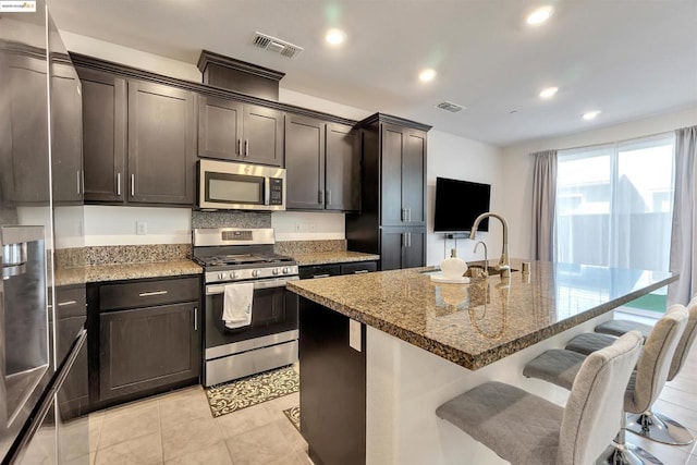 kitchen featuring stainless steel appliances, a kitchen breakfast bar, a kitchen island with sink, light stone counters, and dark brown cabinets