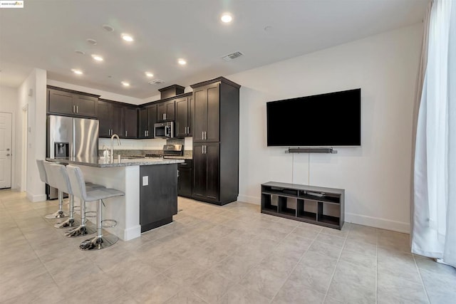 kitchen featuring an island with sink, a breakfast bar area, stainless steel appliances, dark brown cabinets, and light stone counters