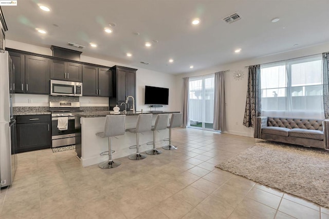 kitchen with stainless steel appliances, dark stone countertops, a kitchen breakfast bar, light tile patterned floors, and a center island with sink