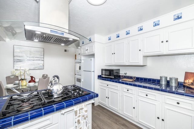 kitchen with white appliances, dark wood-type flooring, white cabinetry, island range hood, and tile countertops