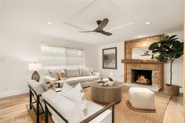 living room with a stone fireplace, a tray ceiling, ceiling fan, and light hardwood / wood-style flooring