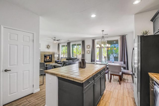 kitchen featuring stainless steel fridge, tile counters, hanging light fixtures, ceiling fan with notable chandelier, and a tile fireplace