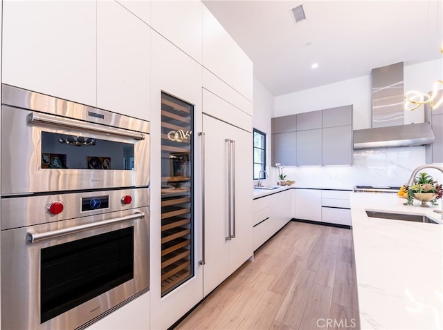 kitchen featuring stainless steel double oven, backsplash, wall chimney range hood, light stone counters, and sink