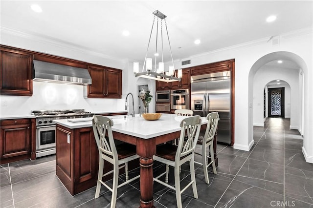 kitchen with pendant lighting, sink, built in appliances, a kitchen island with sink, and wall chimney exhaust hood