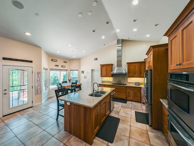 kitchen with appliances with stainless steel finishes, an island with sink, sink, light stone counters, and wall chimney range hood