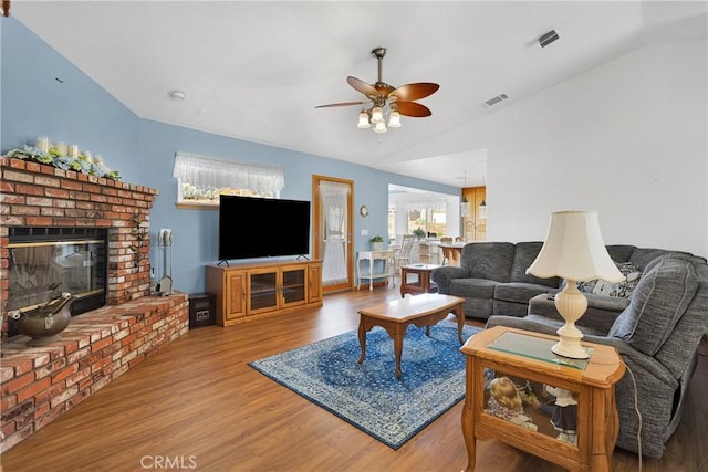 living area with wood finished floors, visible vents, a ceiling fan, vaulted ceiling, and a brick fireplace