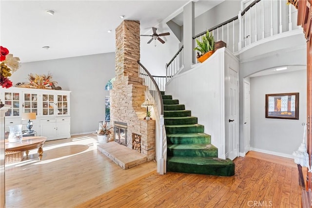 staircase featuring ceiling fan, a towering ceiling, hardwood / wood-style floors, and a stone fireplace