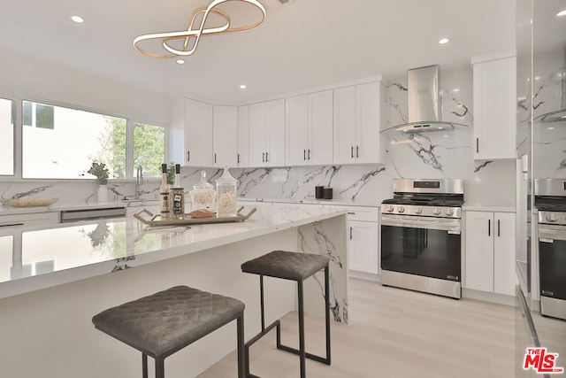 kitchen with white cabinetry, a kitchen breakfast bar, light stone countertops, stainless steel stove, and wall chimney exhaust hood