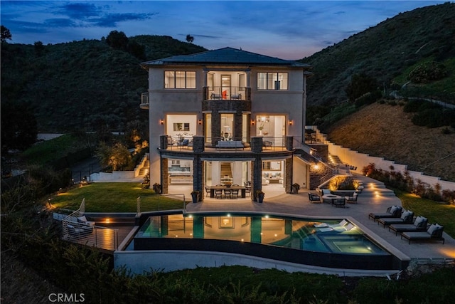 back house at dusk featuring a swimming pool with hot tub, a balcony, and a patio