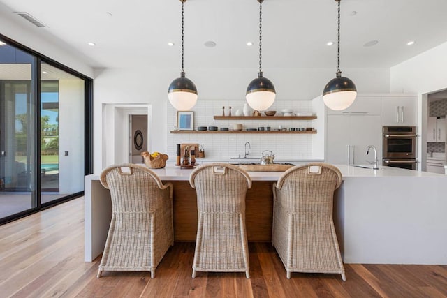 bar featuring stainless steel double oven, white cabinetry, backsplash, hanging light fixtures, and hardwood / wood-style flooring