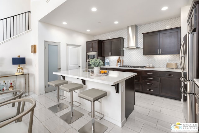 kitchen featuring wall chimney range hood, a kitchen bar, a kitchen island with sink, stainless steel appliances, and dark brown cabinets