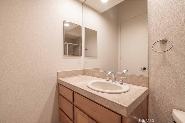 bathroom featuring a shower, a textured wall, vanity, and toilet