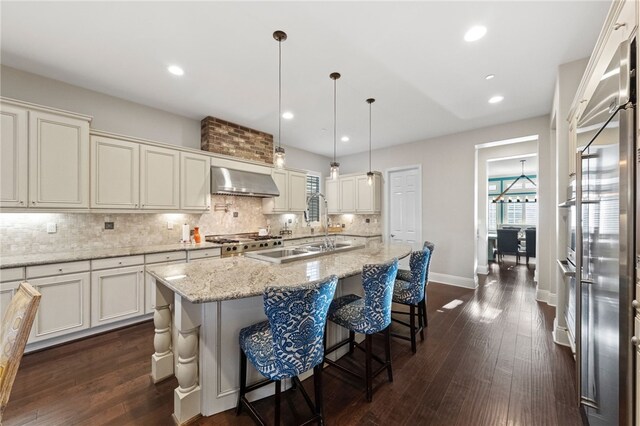 kitchen featuring wall chimney exhaust hood, a kitchen bar, decorative light fixtures, a kitchen island with sink, and light stone counters