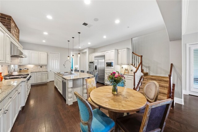 kitchen with pendant lighting, built in appliances, an island with sink, sink, and light stone counters