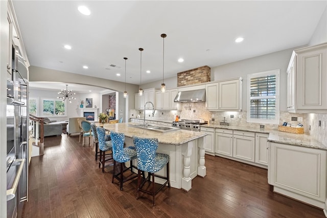 kitchen with light stone countertops, wall chimney range hood, hanging light fixtures, a kitchen island with sink, and a breakfast bar