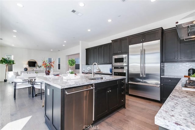 kitchen featuring tasteful backsplash, sink, built in appliances, a kitchen island with sink, and light stone countertops