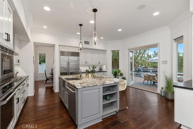 kitchen featuring built in appliances, hanging light fixtures, a kitchen island with sink, sink, and light stone counters