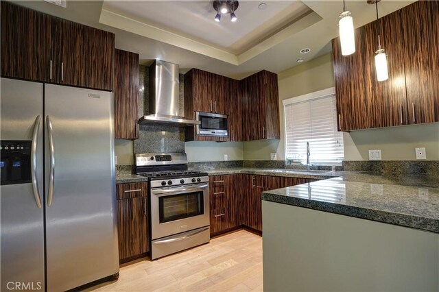 kitchen featuring dark stone countertops, pendant lighting, a raised ceiling, appliances with stainless steel finishes, and wall chimney exhaust hood