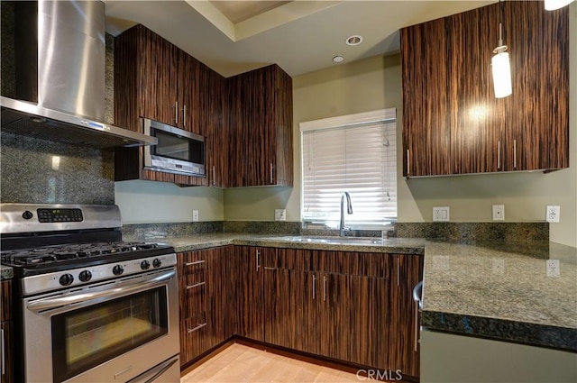 kitchen with stainless steel appliances, dark brown cabinets, dark stone counters, wall chimney range hood, and sink