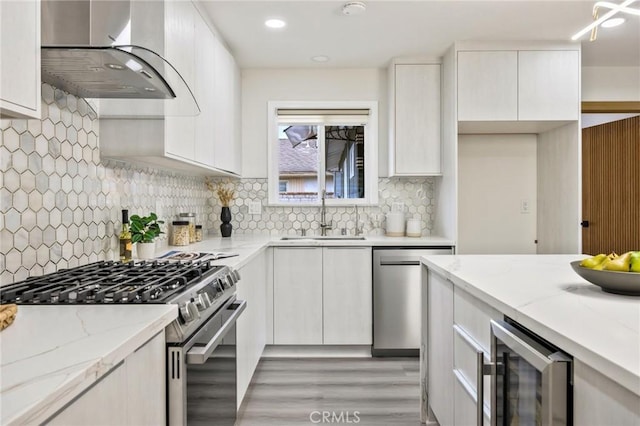 kitchen featuring wine cooler, wall chimney range hood, sink, appliances with stainless steel finishes, and light stone counters