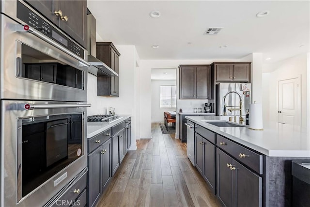 kitchen featuring a center island with sink, appliances with stainless steel finishes, dark brown cabinets, wood-type flooring, and sink