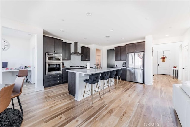 kitchen with stainless steel appliances, wall chimney exhaust hood, a center island with sink, and dark brown cabinetry