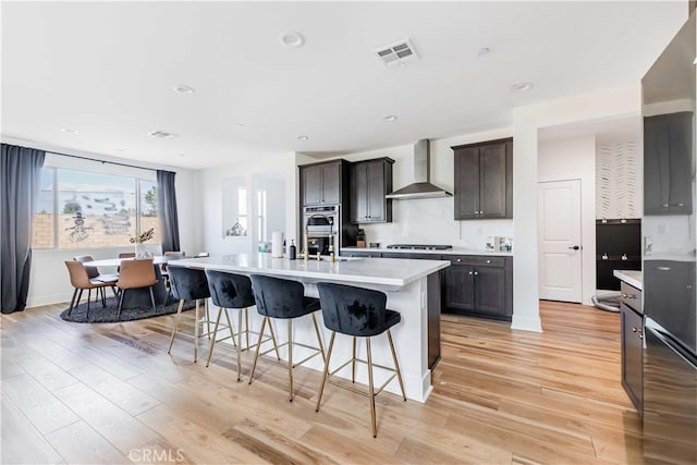 kitchen featuring dark brown cabinets, an island with sink, light hardwood / wood-style flooring, and wall chimney range hood