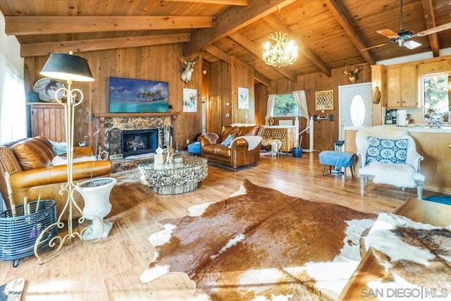 living room featuring a stone fireplace, wooden walls, light wood-type flooring, ceiling fan with notable chandelier, and lofted ceiling with beams