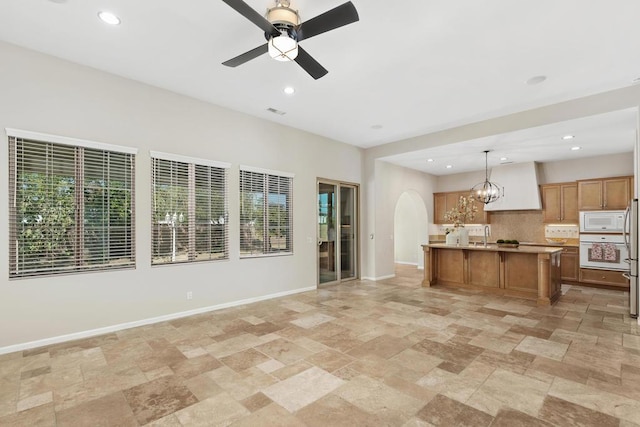 kitchen with a center island with sink, decorative backsplash, white appliances, pendant lighting, and ceiling fan with notable chandelier