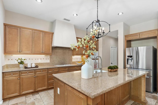 kitchen featuring stainless steel fridge, backsplash, a kitchen island with sink, custom range hood, and cooktop