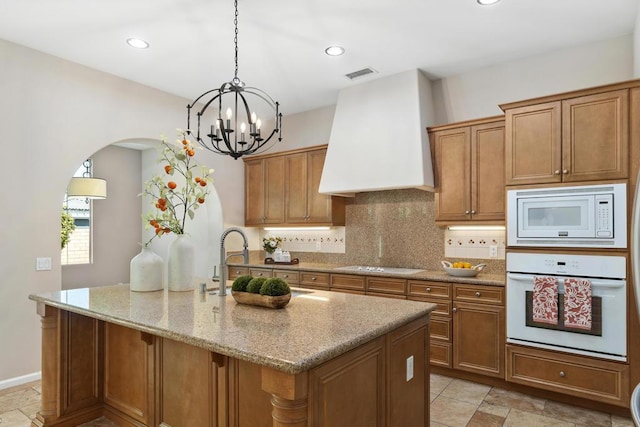 kitchen with a kitchen island with sink, white appliances, hanging light fixtures, custom range hood, and light stone counters