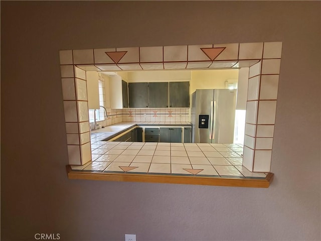 kitchen featuring tile counters, kitchen peninsula, stainless steel fridge, decorative backsplash, and sink